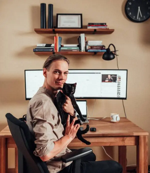 Luka Šolta drži crnu mačku i sjedi u kompjuterskoj stolici s drvenim stolom i širokim monitorom u pozadini